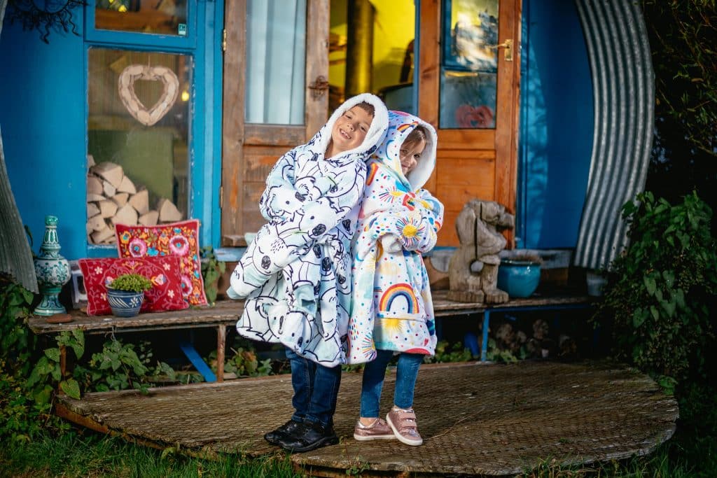 Boy in koala cwtchie and girl in rainbow cwtchie blanket hoodie outside glamping pod