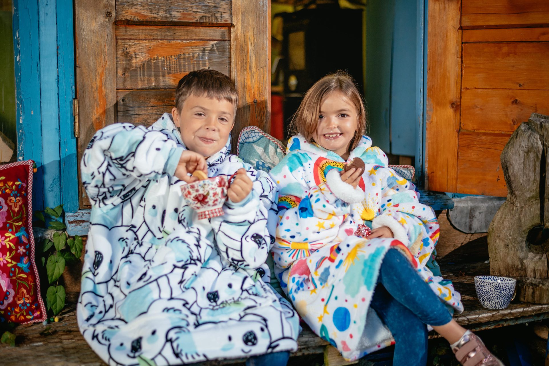 Boy in koala and girl in rainbow hoodie blanket drinking hot chocolate in front of glamping pod